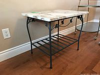 Side view of rectangular marble top side table with decorative black metal base and lower slatted shelf on hardwood flooring near wall.