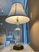 Full view of the lamp showing clear glass base with diamond pattern, brass base, and white fabric shade on a marble top table near window.