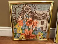 Front view of one framed needlepoint showing house, flowers, and tree in pastel color needlepoint.