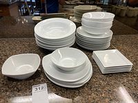 Overall view of stacked white plates and bowls on a granite countertop showing all types included in the lot