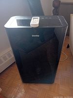 Front view of the black Danby portable air conditioner with remote control placed on top and the exhaust hose attached at the back.