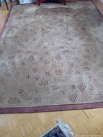 Full view of the patterned carpet on wood floor, showing beige background with reddish-brown abstract shapes and leaf designs, solid colored border