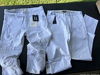 Two white pants laid flat, showing one pair with distressed details and both with tags attached.