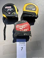 Three tape measures placed on a flat surface: Tool Bench Hardware, Stanley, and Milwaukee brands visible.