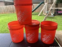 Four orange plastic buckets with Home Depot logo, one bucket stacked on another. Bright outdoor setting showing all buckets in good shape.