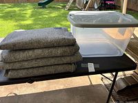 Four gray moving blankets stacked next to a large clear plastic tote with gray lid on a table outdoors.