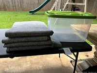 Four gray moving blankets stacked next to a large clear plastic tote with green lid on black table outdoors.