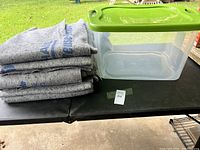 Full view of five folded grey moving blankets stacked beside a clear plastic storage bin with a green lid on a black surface outdoors.