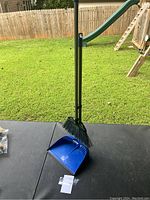 Blue dust pan and black broom standing on dark surface outdoors with grass and fence background.