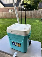 Full view of turquoise and white Igloo cooler with wheels and telescoping handle outdoors on a table.