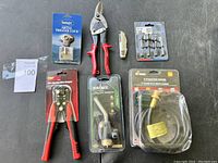 Lot of new tools including propane hose assembly, wire stripper, utility torch, metal trigger lock, tin snips, utility knife, and drill bits arranged on table.