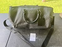Full view of Michael Kors duffle bag resting on a flat surface with grass background. Shows black leather with signature logo pattern, handles, shoulder strap, and zipper.