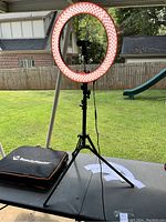 Fully assembled BeautyMaster LED ring light illuminated on black adjustable tripod stand with phone holder, set up outdoors on a table.