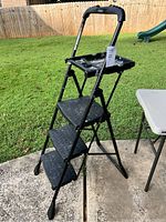 Side-angle view of the black 3-step folding step ladder standing on concrete outdoors.