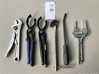 View of six hand tools including wrenches, pliers, and pry bar laid out on table.