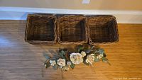 Three square wicker baskets placed on a floor against a wall, along with a faux floral festoon with white flowers and green leaves.