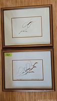 Pair of two framed and matted prints on a wall, one above the other, each with visible brown and wooden framing.