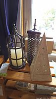 Three candle holders displayed together on a wooden surface by a window: 1) Metal bird cage style holding a white pillar candle, 2) Metal cylindrical lattice design, 3) Ceramic pyramid shaped with star cutouts.