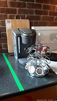 Photo showing Keurig coffee maker on counter, metal coffee pod holder filled with pods, and wooden cutting board behind