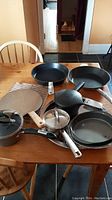 Photo shows two black frying pans, three metal pots (one with glass lid), two racks, large round spatula, round cake pan, and serving platter on wooden table.