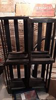 Four black painted wooden crates stacked in two rows of two. Open slat design visible. Background shows basement with exposed brick and some posters.