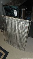 Two metal dog cage panels stacked against kitchen appliances and a trash can. Shows the folded latch clasp on one panel and the grid wire structure of the panels.