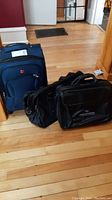 Three luggage pieces: SwissGear blue wheeled weekender and two black leather carry cases on wood floor.