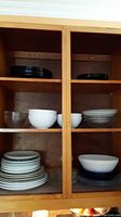 Wide view of several stacks of plates and bowls inside a wooden cabinet. Visible are white ceramic plates, glass bowls, and dark plates, arranged on shelves.