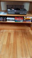 Samsung DVD/VHS player and VHS tapes on wooden shelf, player on upper shelf, tapes below, hardwood floor below shelf visible
