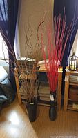 Photo showing two tall dark brown floor vases; one contains natural brown twigs with curls and twists, the other contains straight, painted bright red sticks.