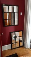 Two window panel-style mirrors, one hung on dark red wall and the other leaning against a white doorframe on a wooden floor.