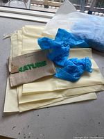 Photo shows four folded cream-colored fabric bags, one cream-colored zipper pouch with 'Nature' embroidered on it, and several blue disposable shoe covers on top.