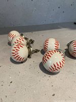 Six white ceramic knobs styled like baseballs, showing red painted stitching details. Metal attachment screws are visible on the underside.