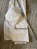 Photo shows folded white fabric shower curtain with silver trim and attached metal grommets, alongside a folded clear plastic shower curtain liner and a metal shower bar placed on a carpeted surface.