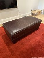 Full view of rectangular brown leather ottoman on red carpet, showing top and sides.