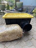 View of the black plastic storage bin shelter with yellow lid and wrapped straw insulation bundle beside it.