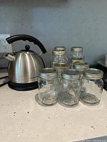 Hamilton Beach stainless steel kettle with black handle and spout alongside nine clear glass mason jars with metal lids, arranged on kitchen counter.