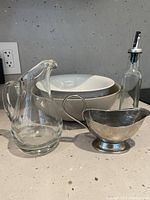 Photo of two ceramic serving bowls (one grey, one white), a clear glass water pitcher, metal gravy boat, and glass oil bottle with pour spout arranged on countertop.
