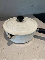 White Le Creuset enamel cast iron saucepan with lid placed on a kitchen counter. Visible concentric rings on the lid and black knob and handle. Shows signs of use.