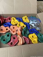 Box containing approximately 20 colorful used rock climbing holds for kids in various shapes and colors.
