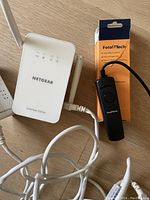 Netgear Essentials Edition Powerline WiFi Extender PLW1010 with LED indicators and antenna connected to cables, alongside the orange box and black wired remote from Foto&Tech.