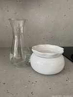 Clear glass vase alongside white ceramic violet pot on countertop with a neutral background.