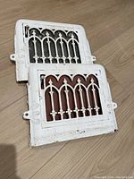Two vintage cast metal heating registers with Gothic arch decorative pattern painted white with visible wear, stacked with one showing red underpaint and the other black underpaint.