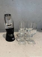 Clear acrylic and black plastic Vinturi wine aerator standing on black no-drip stand next to four clear sherry glasses on countertop.