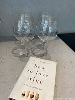 Four clear wine glasses arranged in a square formation with the book 'How to Love Wine' by Eric Asimov in front, on a grey surface.