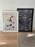 Two wine-themed framed pictures standing on floor, one black frame and one white frame.