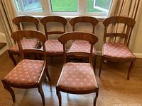 Six wooden dining chairs arranged with pink floral upholstered seat cushions, one chair at front and others staggered behind. Chairs have curved backs and carved legs.