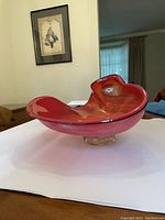Front side view of large red glass bowl with gold flecks on a white surface, showing its irregular, curved shape and clear pedestal base.