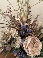 Close-up detail of the artificial flower arrangement showing cream blush peony-like flower, blue imitation berries, and assorted leaves and spikes.