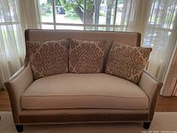 Front view of beige couch with three patterned pillows and nailhead detailing on leather trim.